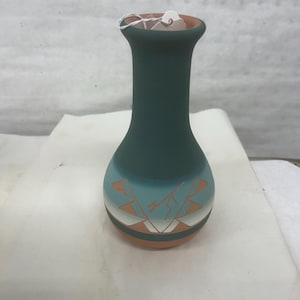 May include: A teal ceramic vase with a narrow neck and rounded body. The vase has a turquoise band with geometric orange and white designs. The rim and base are terracotta. A small tag is attached to the rim.