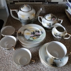 May include: A white porcelain tea set with a Japanese-inspired design. The set includes a teapot, sugar bowl, creamer, and four teacups and saucers. The teacups and saucers feature a delicate floral pattern with a mountain scene.
