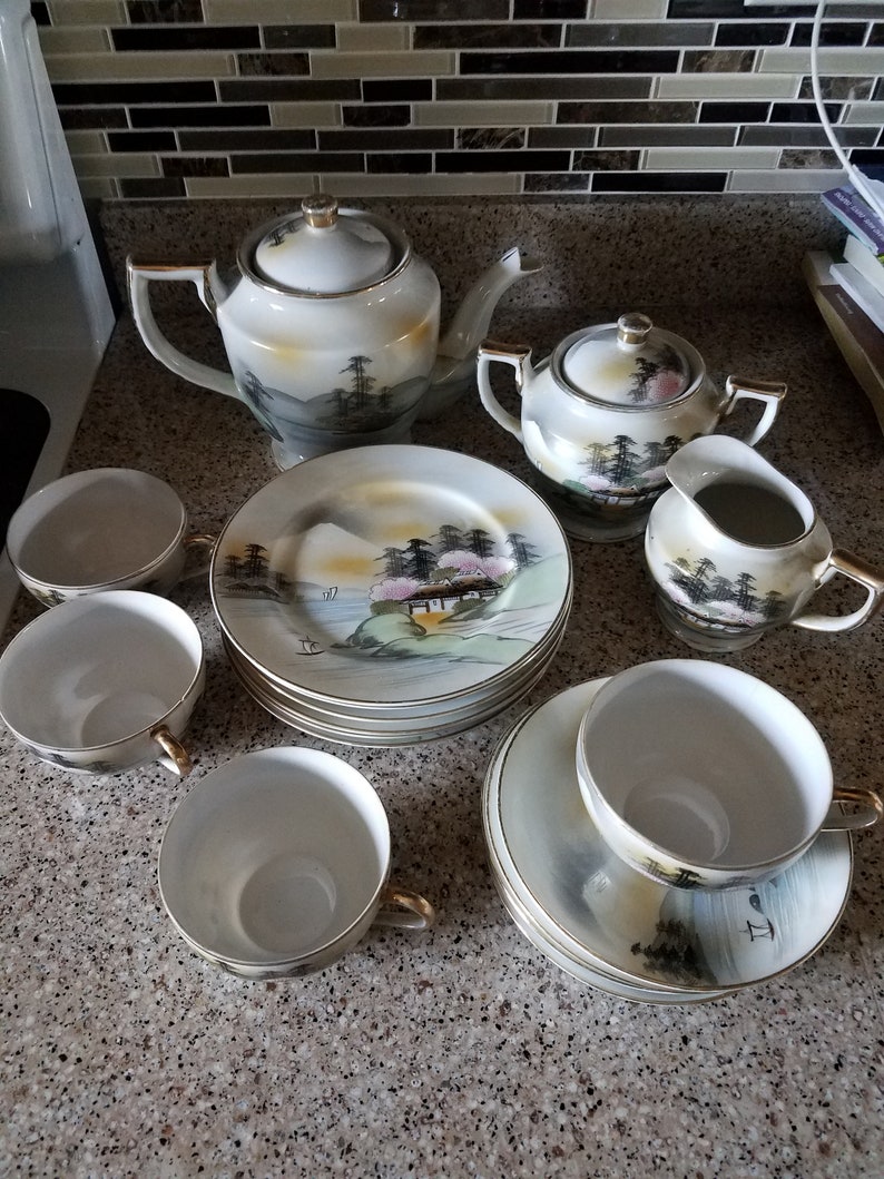 May include: A white porcelain tea set with a Japanese-inspired design. The set includes a teapot, sugar bowl, creamer, and four teacups and saucers. The teacups and saucers feature a delicate floral pattern with a mountain scene.