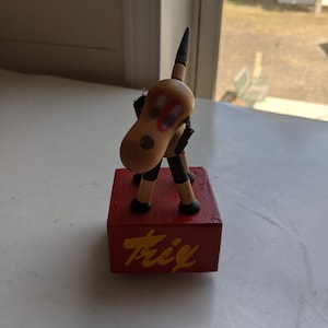 May include: A wooden toy dog with black and white stripes on its legs, red eyes, and a yellow nose. The dog is standing on a red wooden block with the word "Trick" written in yellow letters.