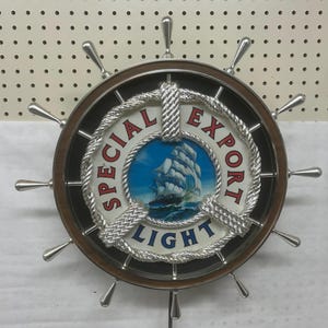 May include: A round, nautical-themed wall decoration resembling a ship's wheel. The center features a painting of a sailboat on the sea, surrounded by the words "SPECIAL EXPORT LIGHT". The wheel is brown with silver accents.
