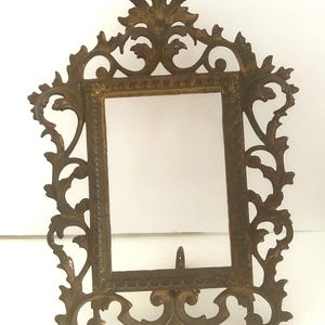 May include: A gold-toned metal picture frame with an ornate, intricate design. The frame has a rectangular opening and a decorative top.