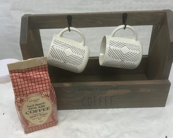 May include: A wooden coffee station with two white mugs featuring a black geometric design, hanging from hooks. A bag of coffee beans sits beside the station. The words "FRESHLY BREWED COFFEE" are printed on the wooden box.