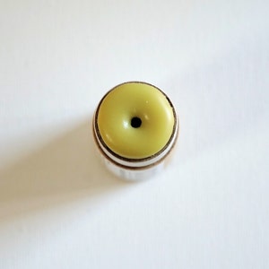 May include: A close-up shot of a small, round object with a light green top and a dark center. The object has a silver-colored metal band around the base and a cork-like material beneath the band. The object is centered on a white background.