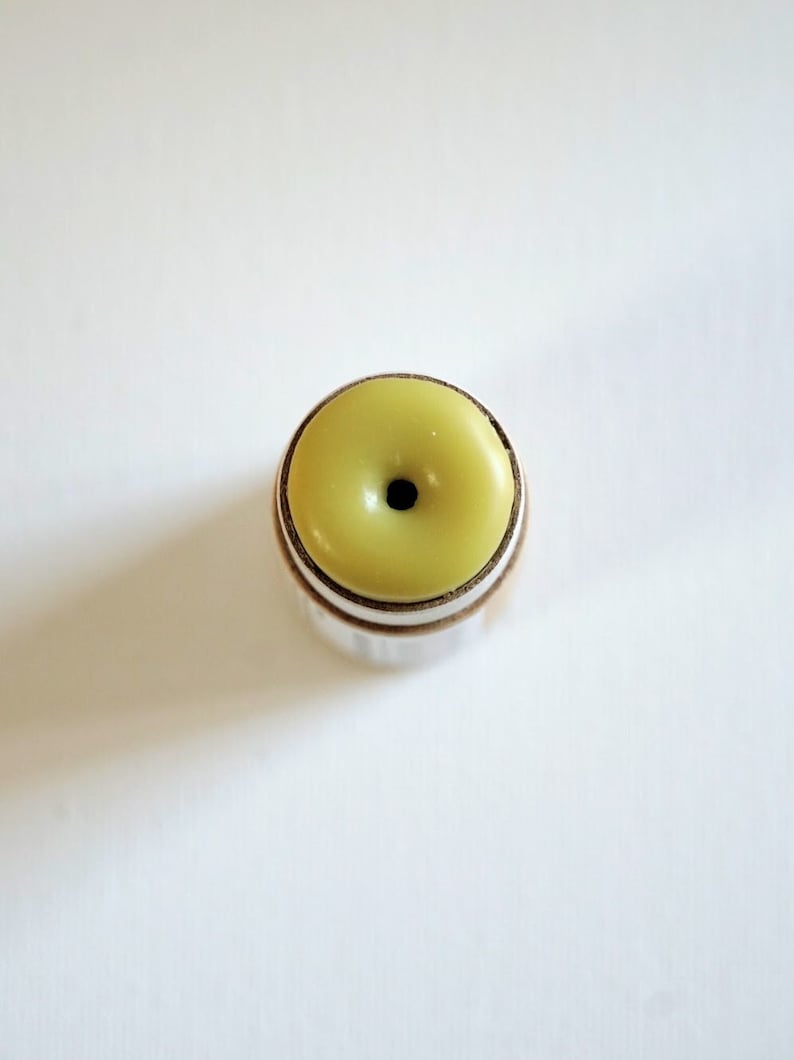 May include: A close-up shot of a small, round object with a light green top and a dark center. The object has a silver-colored metal band around the base and a cork-like material beneath the band. The object is centered on a white background.