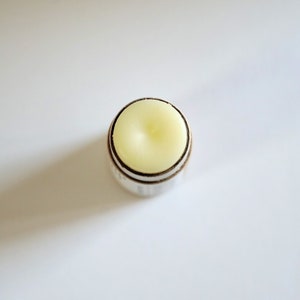 May include: A close-up shot of a lip balm stick. The balm is a pale yellow color, set in a silver and brown container. The top of the balm has a slightly rounded shape. The background is plain white.