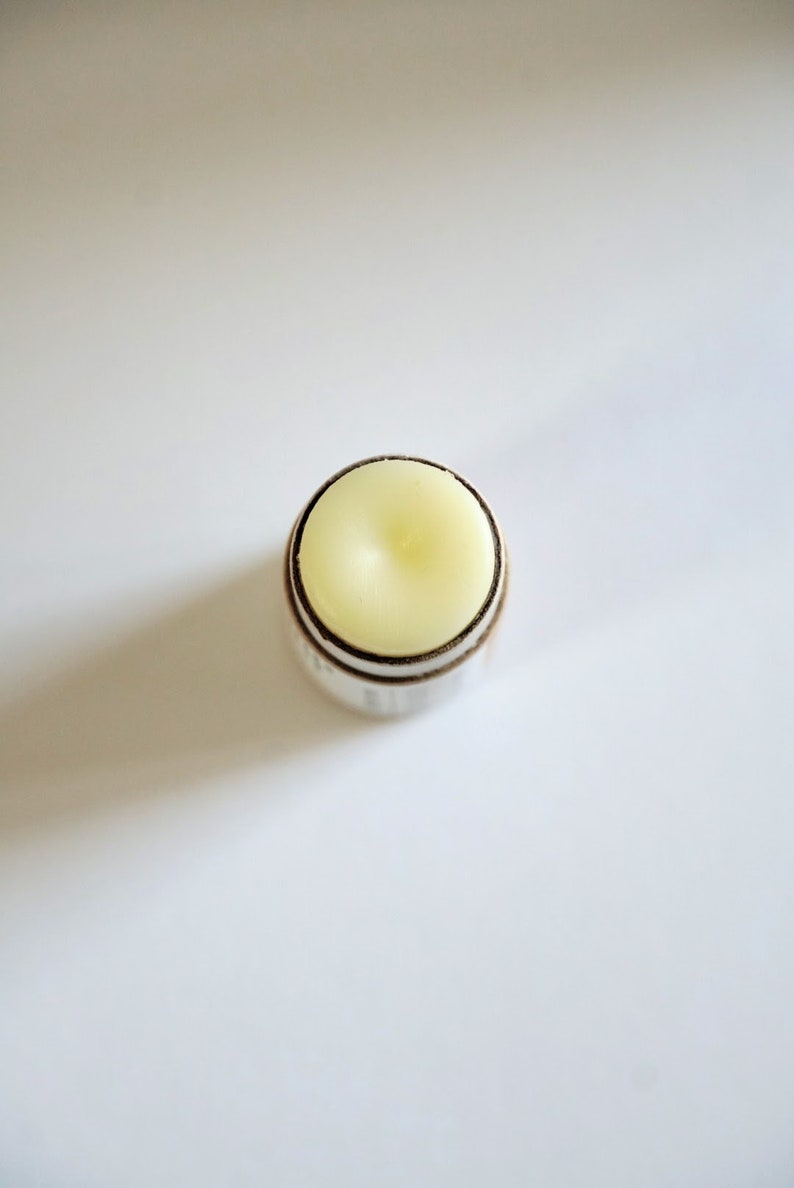 May include: A close-up shot of a lip balm stick. The balm is a pale yellow color, set in a silver and brown container. The top of the balm has a slightly rounded shape. The background is plain white.