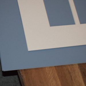 May include: A layered artwork with a light blue mount and a white inner mount with rectangular apertures. The piece is set against a wooden surface, creating a contrast of textures and colours.