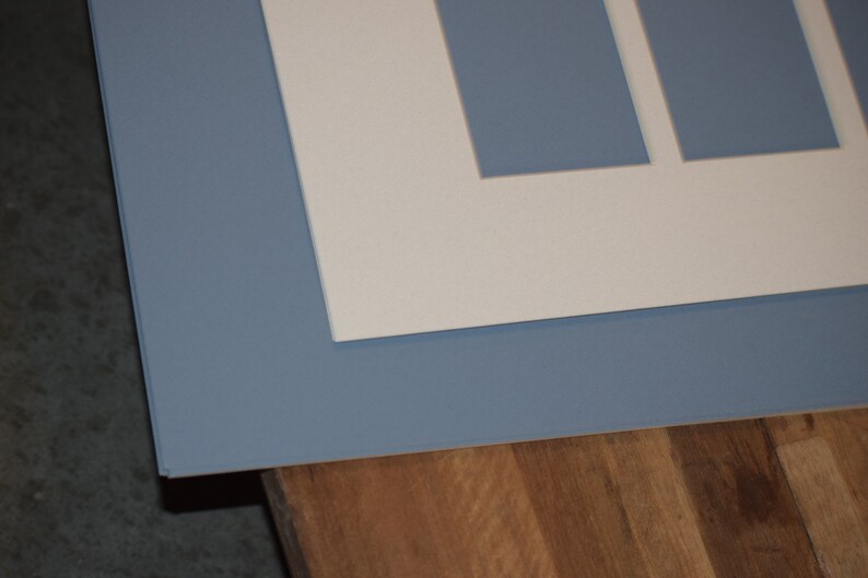 May include: A layered artwork with a light blue mount and a white inner mount with rectangular apertures. The piece is set against a wooden surface, creating a contrast of textures and colours.