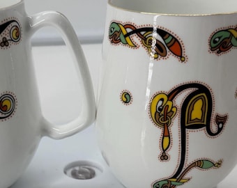 May include: Two white ceramic mugs with decorative Celtic knot designs in black, yellow, blue, and red. Each mug has a handle and a gold rim. The mugs are ideal for coffee or tea.