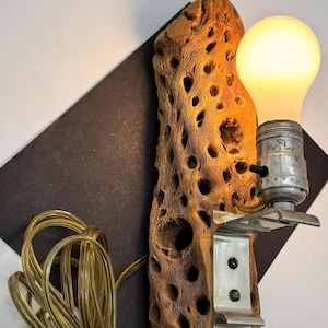 Puede incluir: Un aplique de pared único con una bombilla en un casquillo de metal fijado a un trozo de madera natural y porosa. La madera es de color marrón claro. Un cable eléctrico dorado está enrollado debajo.