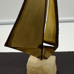 Puede incluir: Una escultura de velero de metal dorado con una base negra y una pequeña bandera. La escultura está sobre una base de mármol blanco. La escultura tiene una etiqueta que dice "Maris Jason Originals".