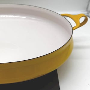 Puede incluir: Una sartén de hierro fundido esmaltado amarillo con interior blanco. La sartén tiene dos asas de bucle y se apoya sobre una superficie negra. Esta batería de cocina es ideal para cocinar.