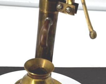 May include: A miniature brass water pump with a detailed design. The pump features a cylindrical body, a curved handle, and a small spout. The base is a tiered, circular design. The pump is a decorative item.