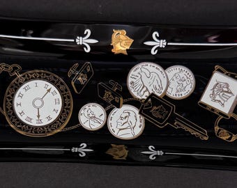 May include: Black rectangular tray with a white border and gold accents. The tray features a clock face, keys, and coins with images of birds and people. The tray has a fleur-de-lis design on each corner.