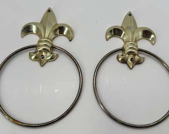 Puede incluir: Dos toalleros decorativos. Cada anillo presenta un diseño de flor de lis dorado en la parte superior, con un anillo circular debajo. Los anillos son de metal y están diseñados para montarse en la pared.