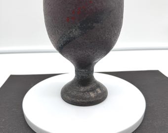 May include: A dark brown, textured ceramic goblet with a speckled finish. The vessel features a wide, rounded bowl tapering to a narrow stem and a circular base. A diagonal black brushstroke accent is visible, complemented by small red dots.
