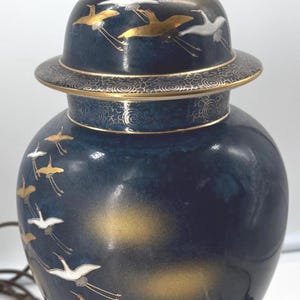 Könnte beinhalten: Dekorative Keramiklampe mit dunkelblauer Glasur, goldenen Akzenten und einem Design fliegender Kraniche. Die Lampe hat eine Messingarmatur und einen dunklen Holzsockel. Ein dekorativer Einrichtungsgegenstand.