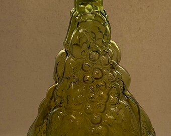 May include: A green glass bottle shaped like a basket of fruit. The bottle has a textured surface and a green glass stopper in the shape of a teardrop.