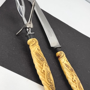 Pode incluir: Um conjunto para esculpir com garfo e faca. As alças são creme com um design esculpido. O garfo tem dois dentes e um topo decorativo. A faca tem uma lâmina longa e prateada.