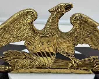 May include: A polished brass eagle emblem featuring a spread-wing design, a central shield with three stars and vertical stripes, and a bundle of arrows held in the talons. The metallic surface has a reflective gold finish with detailed feather textures.