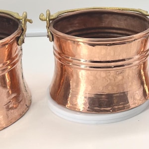 May include: Three copper buckets of varying sizes with brass handles. The buckets have a hammered texture and a shiny, reflective surface. The buckets are arranged in a row, showcasing their different dimensions and design.