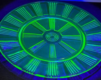 May include: A green glass plate with a wheel design. The plate has a raised rim and a center circle with concentric circles.