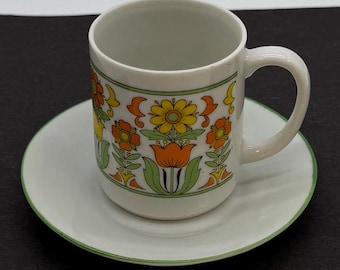 May include: A white ceramic coffee cup and saucer with a green, orange, and yellow floral design. The cup has a rounded handle and the saucer has a green rim.