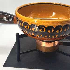 May include: A round ceramic fondue pot with a glossy mustard yellow interior and a dark brown handle. The exterior features a repeating black swirl pattern. The pot rests on a black metal stand with a central copper-coloured burner component.
