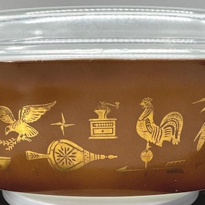 Puede incluir: Una fuente para horno Pyrex marrón con tapa de cristal transparente. La fuente presenta ilustraciones doradas de varios objetos, como un gallo, un águila y un molinillo de café. La fuente está sobre una superficie blanca.