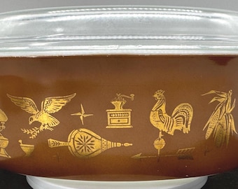 May include: A brown Pyrex casserole dish with a clear glass lid. The dish features gold-colored illustrations of various objects, including a rooster, eagle, and coffee grinder. The dish is sitting on a white surface.