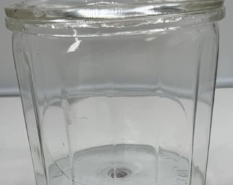 May include: A clear glass canister with a matching lid featuring a knob handle. The body of the container has vertical panels and embossed text at the base that reads EXCLOBA 115. The glass is transparent, showing a smooth, multi-faceted design.