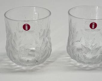 May include: A set of four clear glass tumblers featuring a textured botanical pattern of leaves and stems. Each glass has a circular red sticker with a white letter i and the text IITTALA MADE IN FINLAND printed on the front surface of the glass.