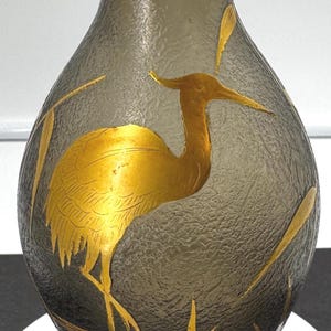May include: A decorative glass vase with a textured surface and a gold rim. The vase features a gold heron design and gold accents. The base is black with a gold trim. A unique home decor item.