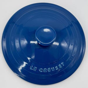 Pode incluir: Uma tampa redonda de ferro fundido esmaltado em azul profundo com um puxador central e sulcos circulares concêntricos. O nome da marca LE CREUSET está gravado em letras maiúsculas ao longo da borda inferior da tampa de acabamento brilhante.