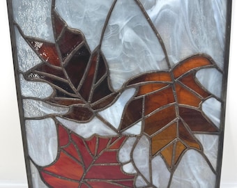 May include: A rectangular stained glass panel featuring three autumn leaves in shades of deep red, burnt orange, and dark brown. The leaves are set against a background of swirled, translucent white glass, all held together by dark metal cames.