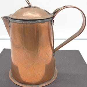 May include: A copper-colored metal coffee pot with a lid and handle. The pot has a spout and a rounded base. The handle is attached to the side of the pot. The lid has a small loop on top.