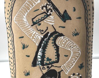 May include: A tan ceramic bottle with a painted folk art design. The design features a dancing figure in black pants, a green vest, and a white shirt, holding a tool. The bottle has a textured surface and green accents.