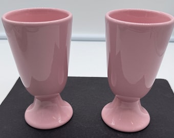 May include: Two pink ceramic cups with a pedestal base. The cups are empty and have a smooth, glossy finish.