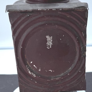 May include: A vintage, maroon-colored lantern with a square body and a cylindrical top. The lantern has a decorative, ridged design and a small, round base. The electrical components are visible at the bottom. The lantern is likely made of metal.