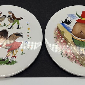 May include: Two white ceramic plates with colorful illustrations. One plate features a rooster-like figure with a sheep and people gathering. The other plate depicts a character with a red hat and green jacket. Floral and bird accents.