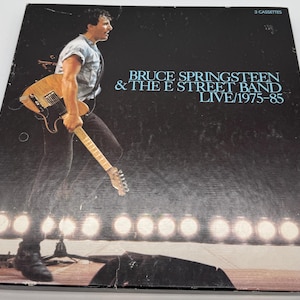 Puede incluir: Una portada de álbum en blanco y negro con una foto de Bruce Springsteen tocando la guitarra. El título del álbum es "Bruce Springsteen & The E Street Band Live/1975-85" y el texto "3 Cassettes" está en la esquina superior derecha.