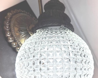 May include: Vintage wall sconce with a clear, textured globe shade. The sconce features a decorative bronze-colored backplate and a dark, ornate fixture. The globe has a square pattern, and the fixture is mounted on a wall.