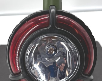 May include: A black and red flashlight with a green handle. The flashlight has a large, round head with a bright light and the text "SMALL SOLDIERS" at the bottom. The handle has a ribbed texture.