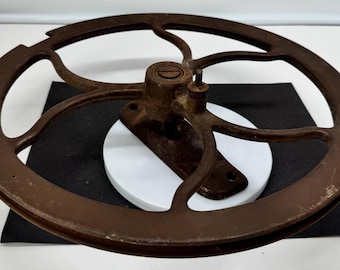 May include: A circular, brown, rusted metal flywheel featuring a central hub with a flat-head screw and a small vertical pin. The wheel is connected to a rectangular mounting base with two circular holes, showing signs of age and oxidation.