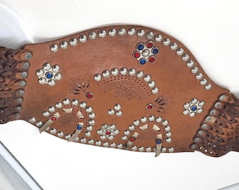 May include: A brown leather horse girth featuring a wide central panel decorated with silver-tone metal studs and inlaid red and blue floral accents. The ends consist of woven leather straps with silver-tone metal buckles for secure fastening.