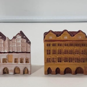 Könnte beinhalten: Vier Miniatur-Gebäudefiguren in einer Reihe angeordnet. Die erste ist ein hoher, metallisch bronzener Turm. Die zweite ist ein hellrosa Gebäude mit Stufengiebel. Die dritte ist ein beiges Gebäude mit Rundbogenfenstern und die vierte ein schmales Haus.