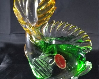 May include: A green and yellow glass sculpture of a fish with a clear glass base. The fish has a label that reads "Murano Made in Italy".