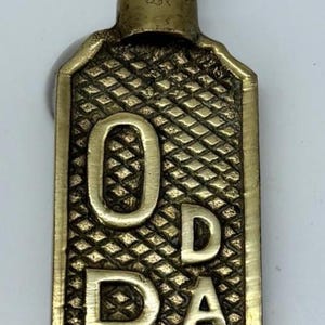 May include: A brass damper control handle featuring a black metal hook at the top. The rectangular brass plate has a textured diamond pattern background and the word "DAMPER" embossed vertically in large capital letters on the front surface.