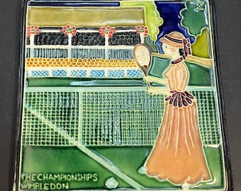 May include: A square ceramic tile featuring a tennis scene at Wimbledon. The tile depicts a woman in a long dress playing tennis, with the text "THE CHAMPIONSHIPS WIMBLEDON" at the bottom. The tile has a black border and a loop for hanging.